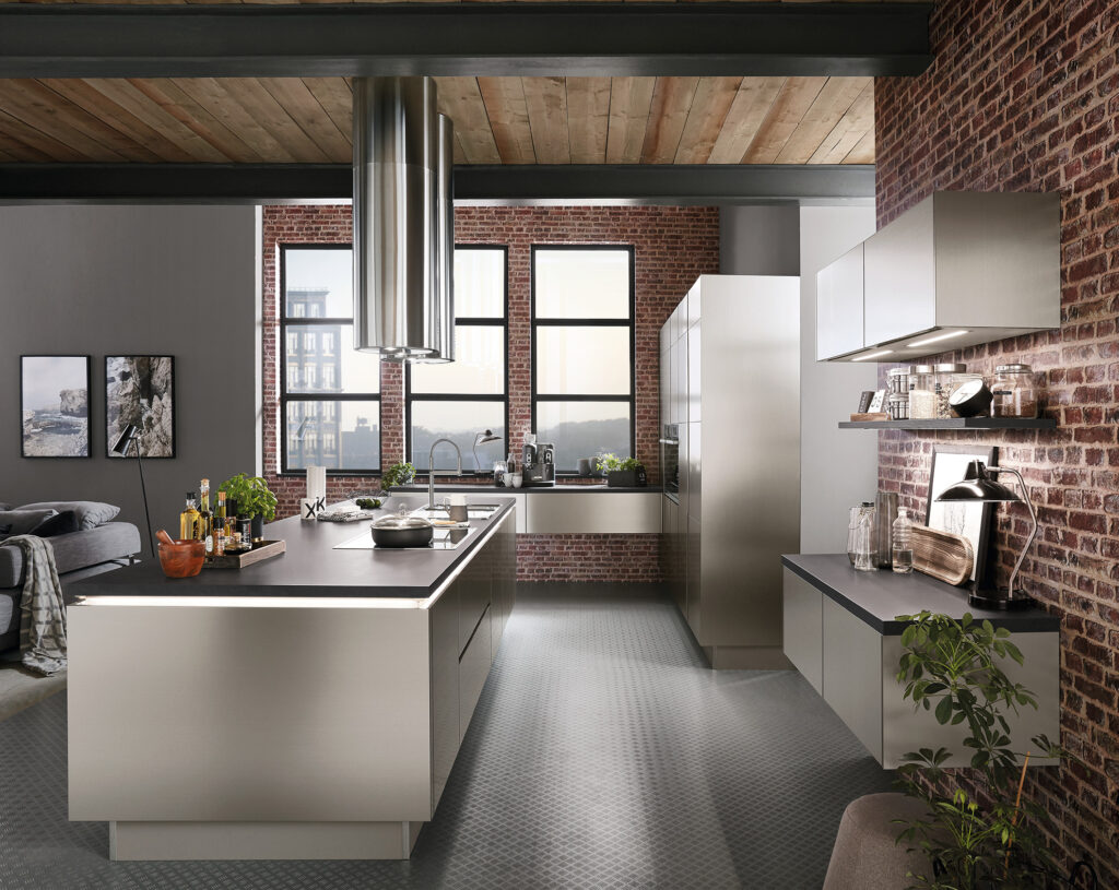 Sleek Iconic Collection kitchen with a central island. The kitchen appears to be in a modern urban loft type home with large windows and exposed brick walls. The cabinets and island are faced with a brushed metal texture, emphasizing the industrial feel of the space.