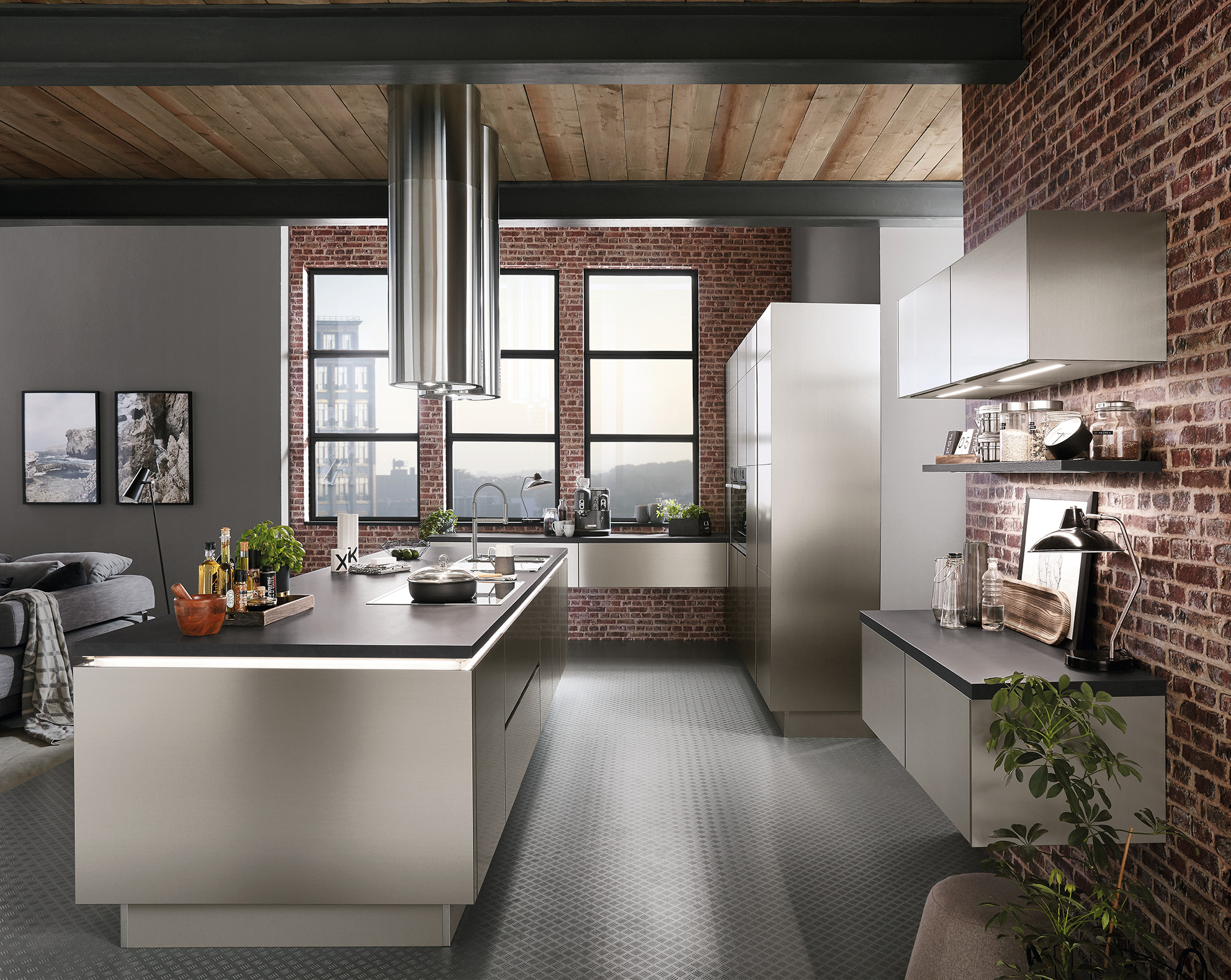 Sleek Iconic Collection kitchen with a central island. The kitchen appears to be in a modern urban loft type home with large windows and exposed brick walls. The cabinets and island are faced with a brushed metal texture, emphasizing the industrial feel of the space.